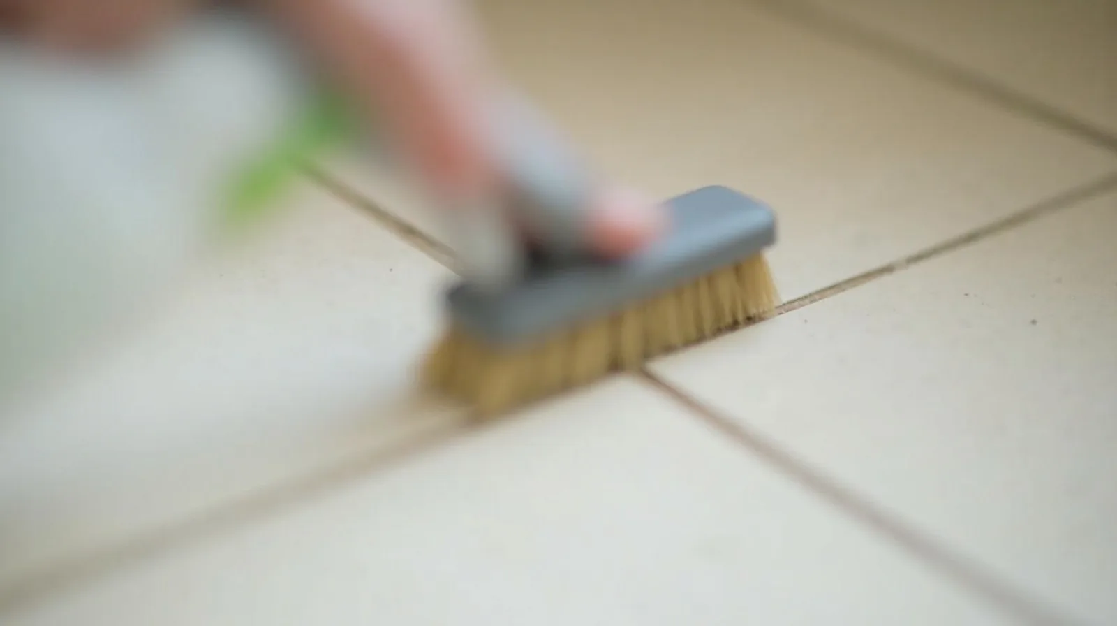 A close-up of a stiff brush scrubbing a bright line of grout between floor tiles, half done